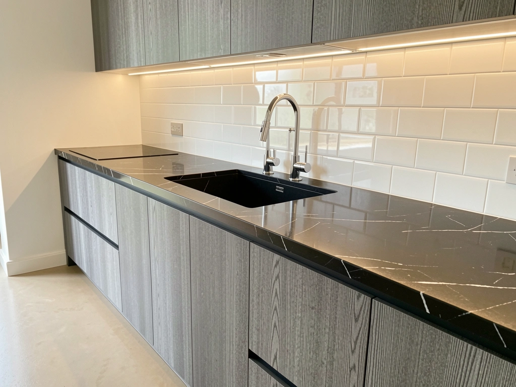 Longa ilha de cozinha com armários cinza escuro texturizados, bancada de mármore preto com veios e pia retangular preta com torneiras cromadas, luz natural refletindo no mármore.