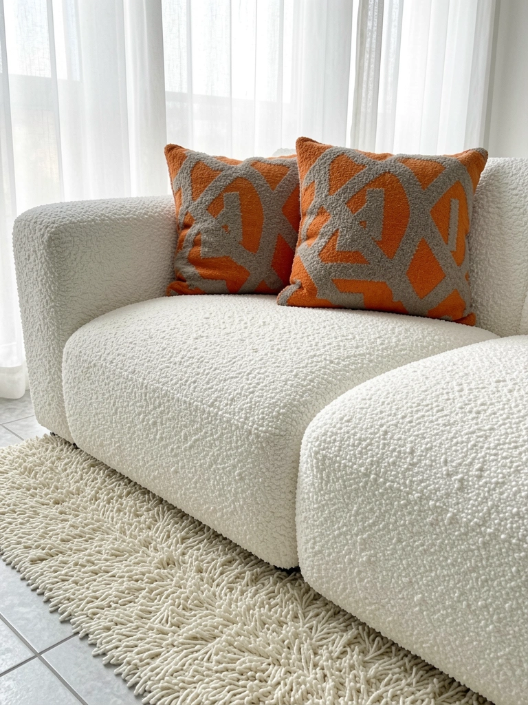 Bouclé branco de sofá modular com almofadas laranja e cinza geométricas, luz natural difusa por cortinas de voal, carpete creme e piso claro.