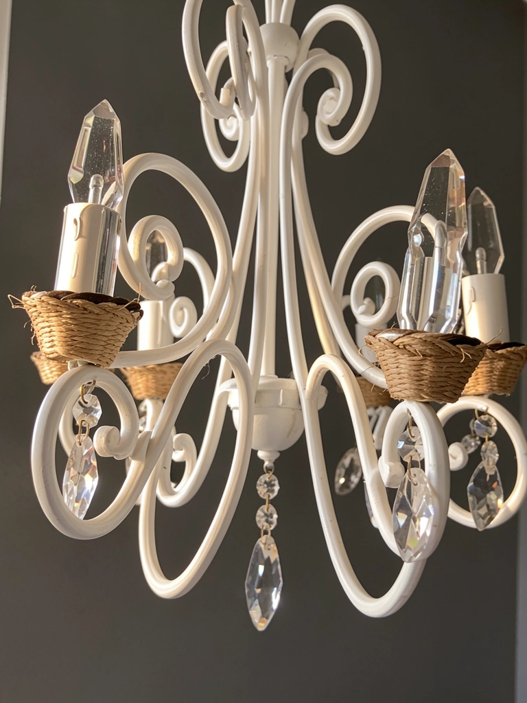Close-up de lustre de ferro forjado branco com detalhes retorcidos, pingentes de cristal e envoltório de fibra natural, montado em parede cinza escuro sob luz natural.