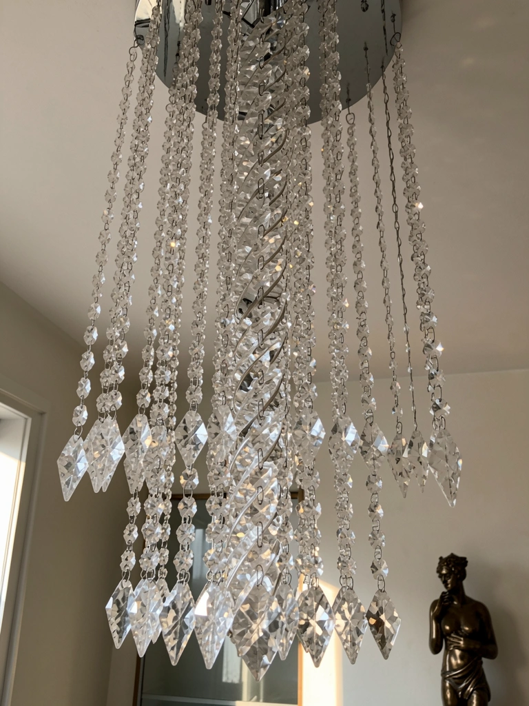 Lustre de cristal moderno com coluna central de cristais em cascata, luz natural refletindo nas superfícies facetadas e escultura de bronze ao fundo.