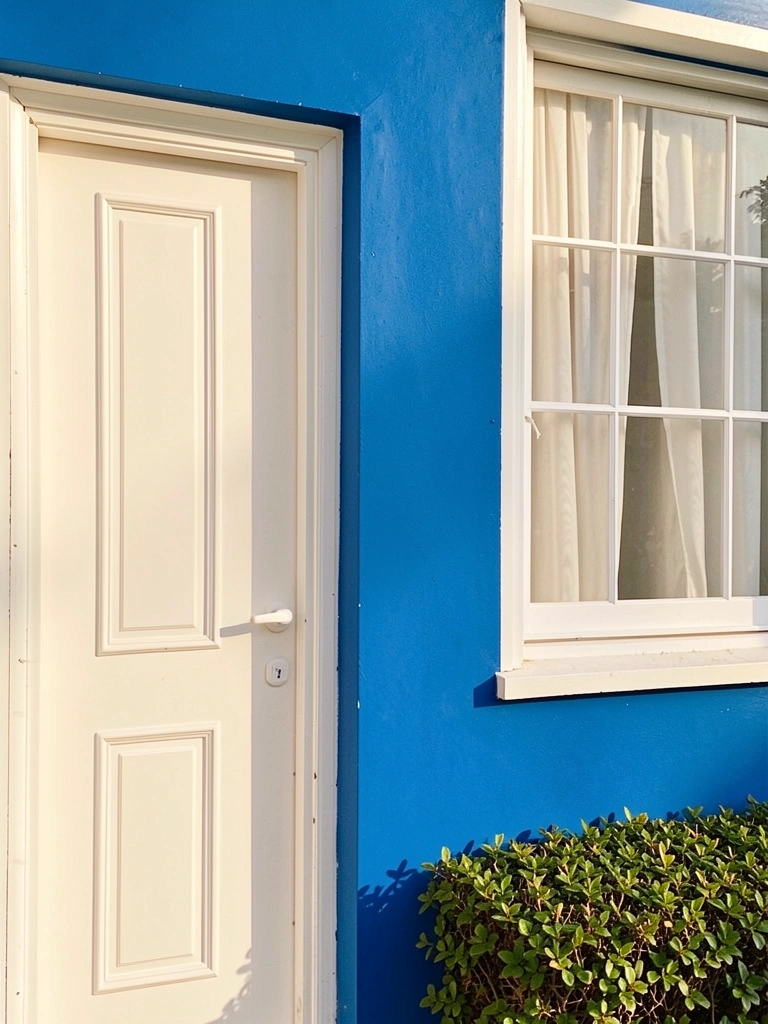 Parede azul vibrante com porta branca de painéis, janela com cortina fluida e sebe verde na base, luz natural com sombras suaves.