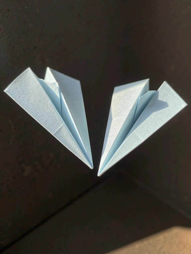 Dois aviões de papel azul-claro suspensos no ar contra fundo escuro, com dobras nítidas e luz natural.