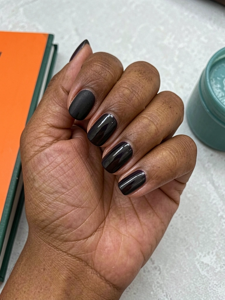 Duas mãos com unhas curtas esmaltadas em tom escuro, uma com acabamento fosco e outra com brilho.