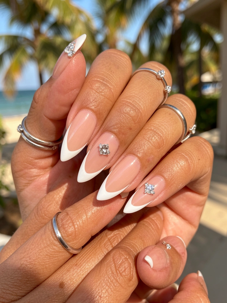 Mãos femininas com unhas francesas alongadas, base nude e pontas brancas, anel prateado e strass brilhante na ponta, luz natural difusa e fundo tropical.