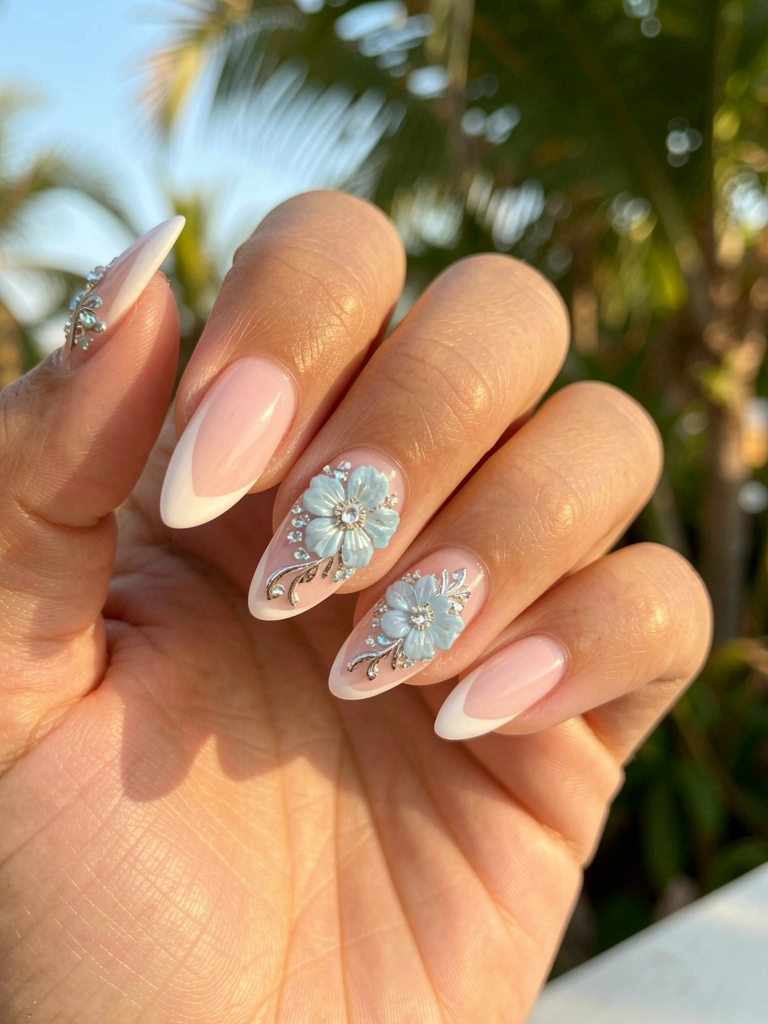 Close-up de mão com unhas francesinhas decoradas com florais azuis e prateados, cristais brilhantes sob luz natural, folhagem tropical ao fundo.