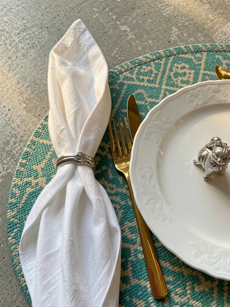 Guardanapo de linho branco com anel prateado sobre esteira teal e bege, prato de porcelana com talheres dourados e gato de prata, fundo cinza com luz natural.