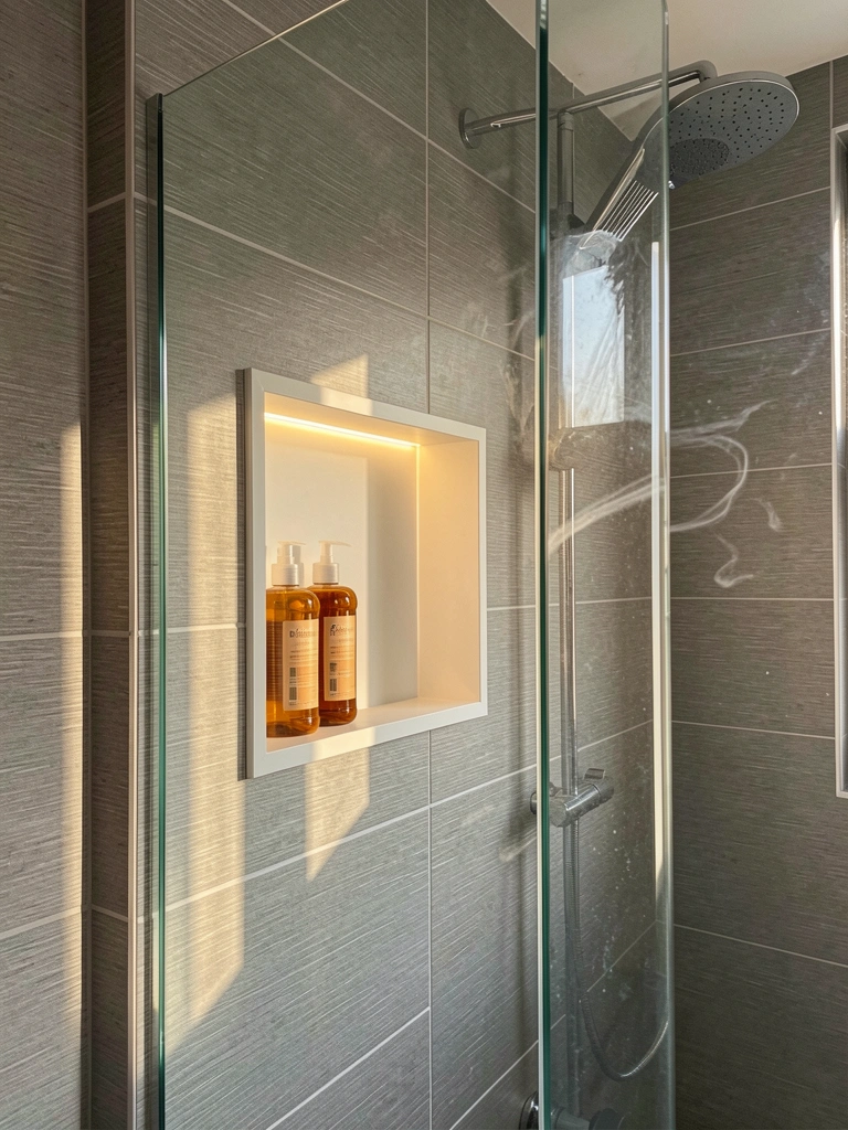 LED ilumina nicho retangular branco com dois frascos âmbar de shampoo e condicionador, paredes de azulejos cinza horizontais e texturizados, box de vidro sem moldura e chuveiro de teto.
