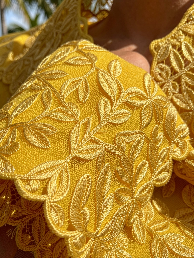 Renda amarela com bordado dourado denso em padrão de folhas, luz natural realçando o brilho dos fios e a textura do tecido.