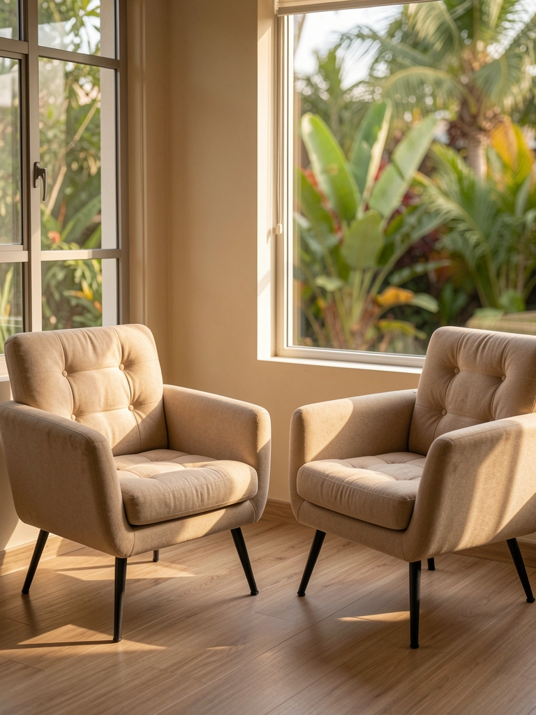 Poltronas estofadas bege com almofadas capitonê e pernas de metal preto, luz natural entrando pela janela, jardim tropical desfocado ao fundo.