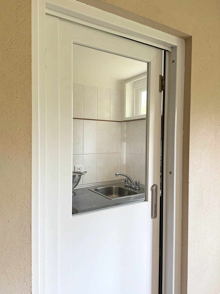Porta de correr branca reflete cozinha com pia de inox, bancada cinza e backsplash de azulejos, luz difusa criando sombras suaves.