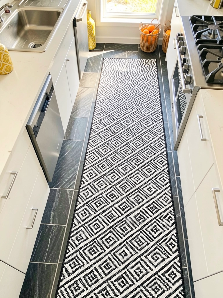 Luz natural incide sobre cozinha com piso geométrico cinza e branco, tapetes xadrez pretos e brancos, cesta de vime com laranjas e garrafa amarela e dourada.