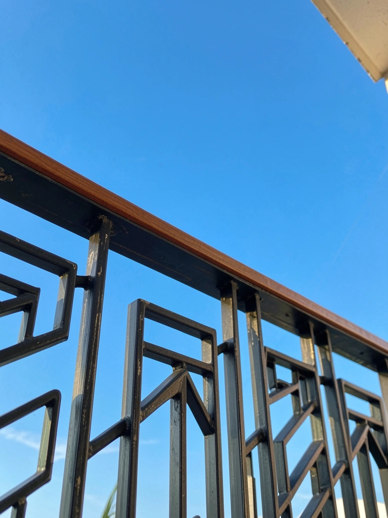 Corrimão de varanda em metal escuro e madeira marrom com padrões geométricos, contra céu azul vibrante sob luz natural.