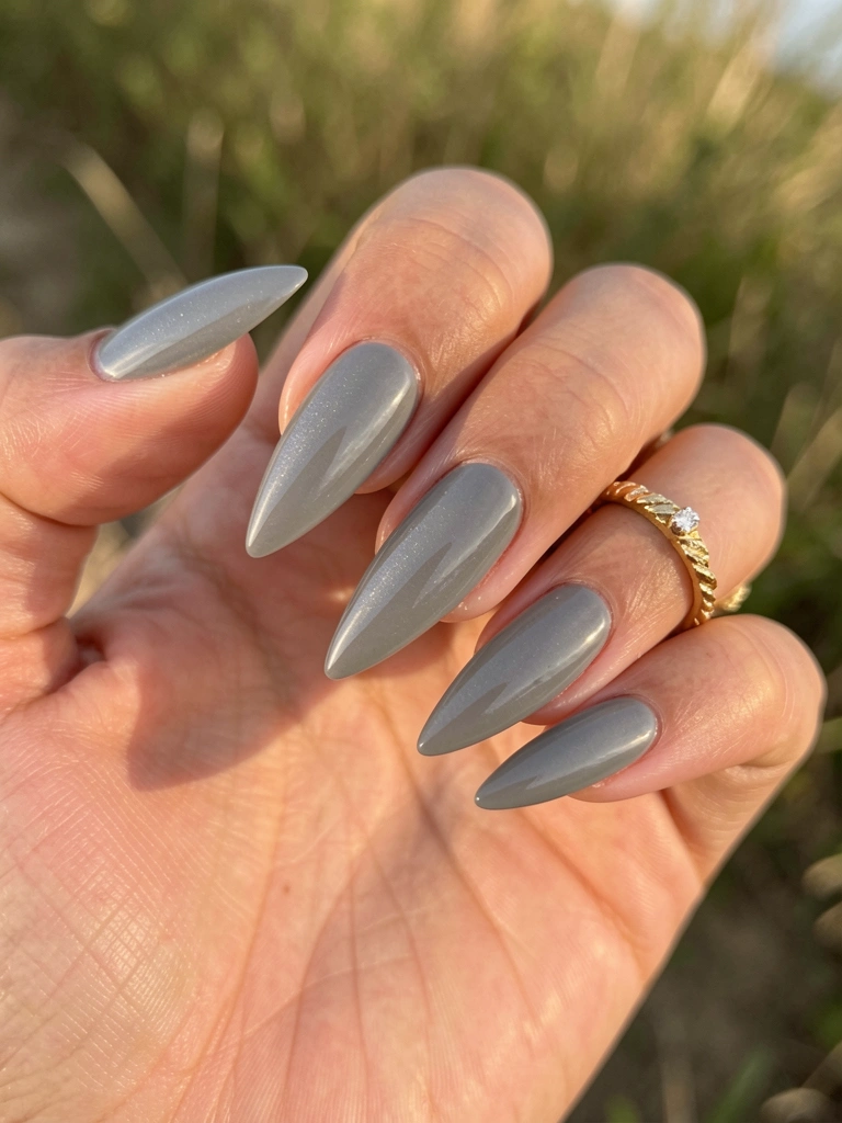 Mão feminina com unhas grisalhas alongadas e pontiagudas, anel de ouro texturizado com diamante, luz natural suave e fundo externo desfocado.