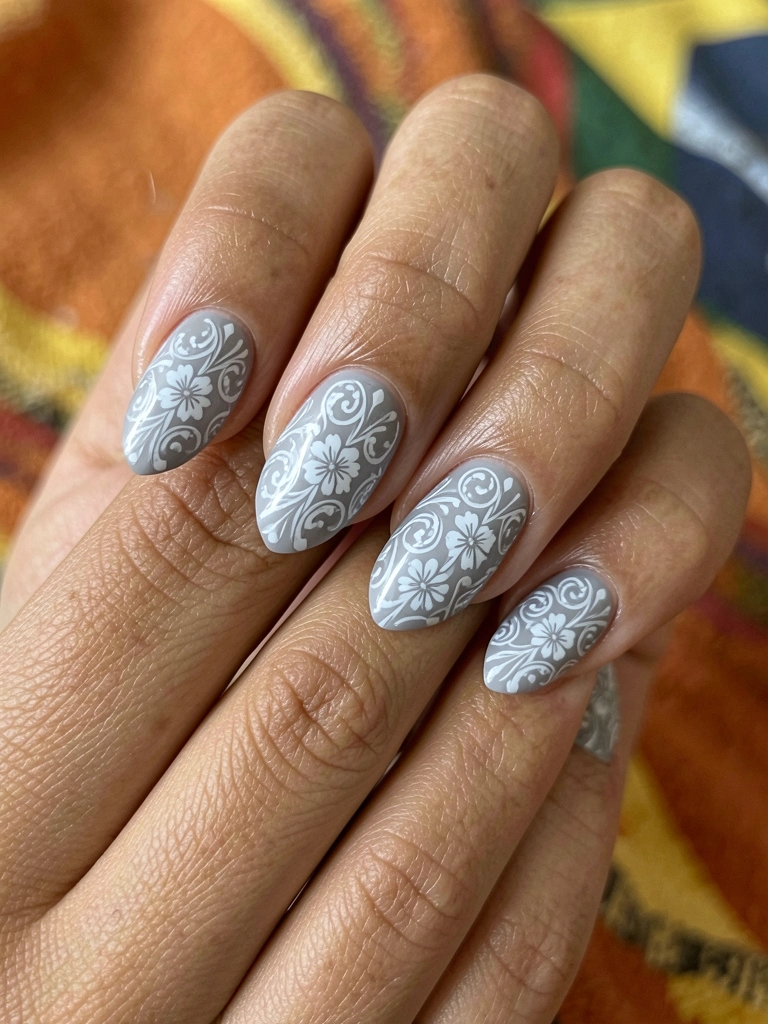 Mãos femininas com unhas cinza e detalhes florais brancos em filigrana, iluminadas por luz natural difusa sobre tecido texturizado.