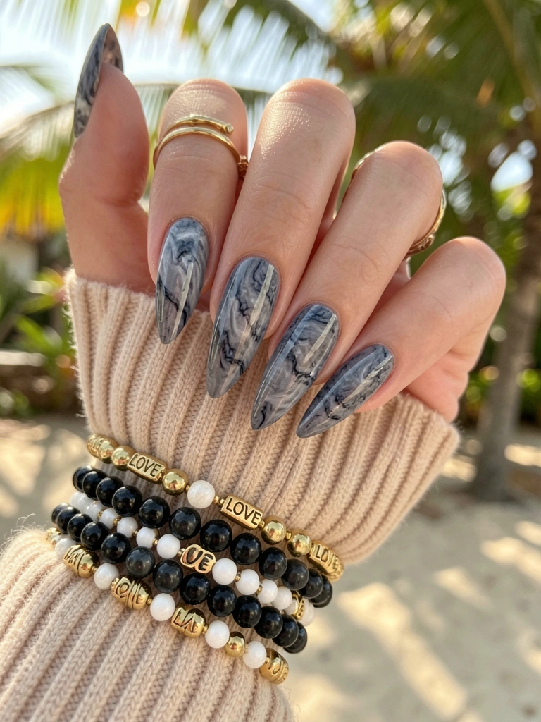 Mãos femininas com unhas em formato amendoado pintadas de esmalte cinza marmorizado, pulseiras de contas pretas, douradas e brancas com pingentes LOVE sobre manga de tricô canelado bege.