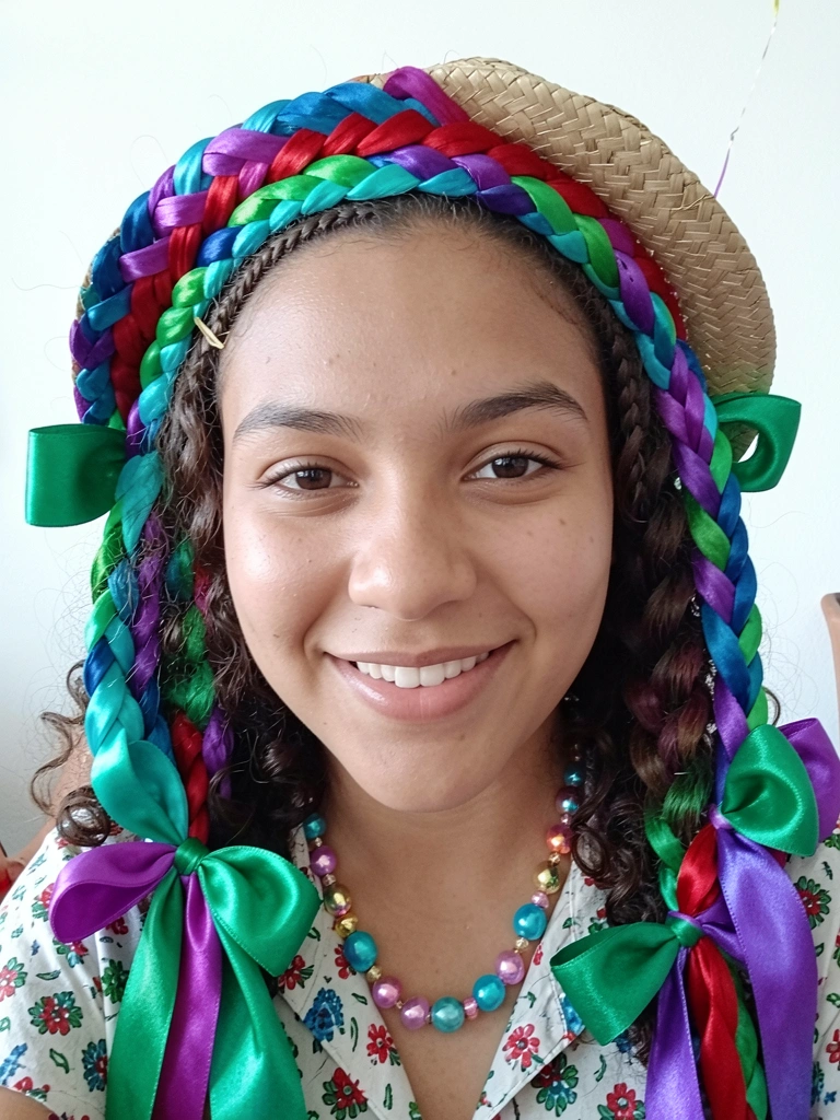Menina com cabelos cacheados e trança lateral embutida, mini chapéu de palha preso na lateral, fundo de festa.