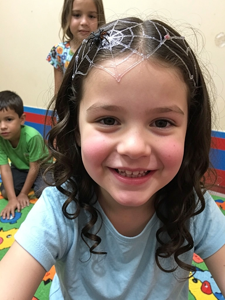 Criança com cabelo cacheado decorado com gel formando uma teia de aranha e uma aranha de brinquedo presa no cabelo.