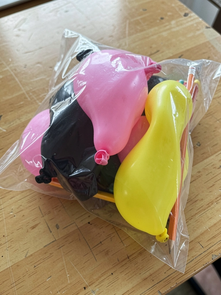 Pacote transparente com balões rosa, preto e amarelo e lápis de papel sobre uma mesa.