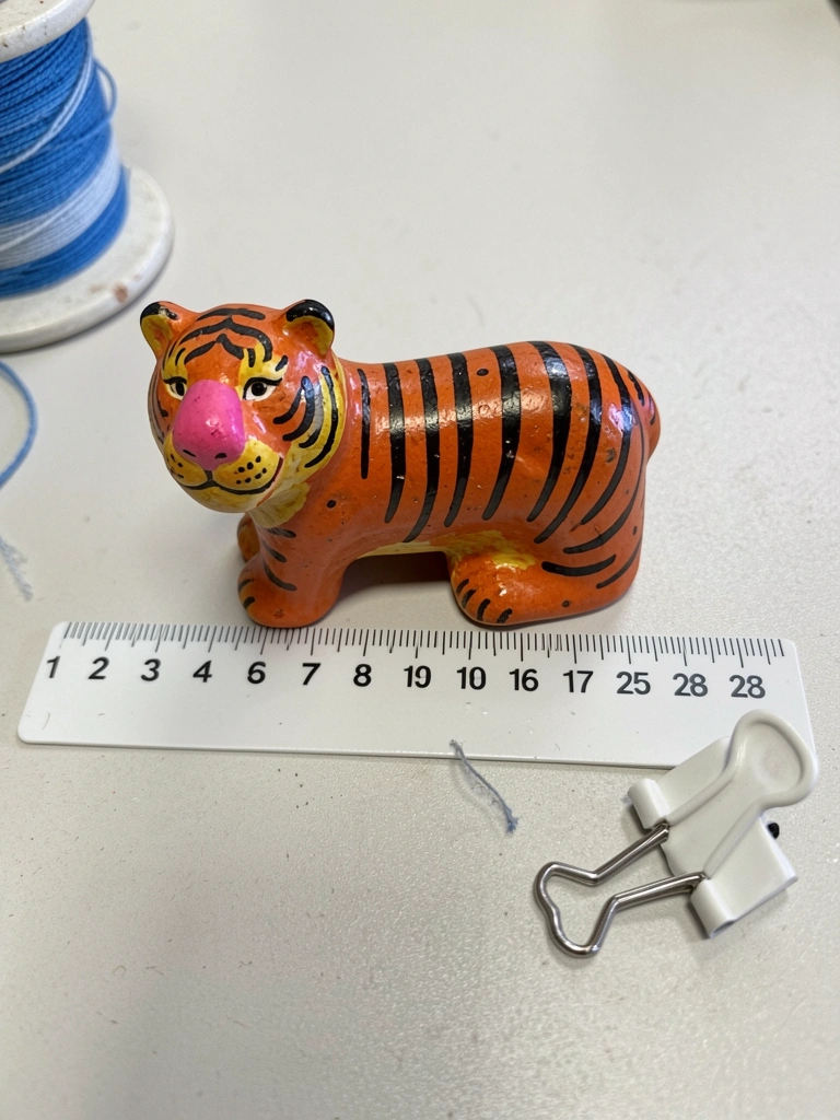 Boneca de biscuit sentada em um laço de cabelo, com mochila mini nas costas e segurando um lápis, fundo claro.