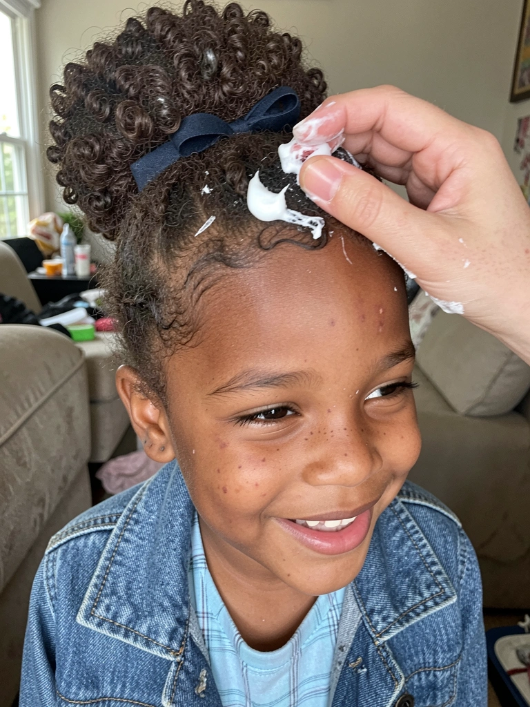 Criança com cabelo cacheado recebendo creme para pentear nas mãos de um adulto.
